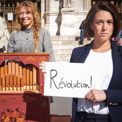 Musiikin vallankumous -sarjan toimittaja Suzy Klein pitelee kylttiä, jossa lukee "Révolution!". Vieressä posetiivari Pariisin kuninkaallisen oopperan portailla.