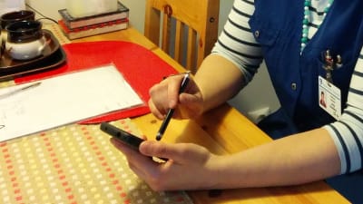 Hemvårdares händer vid ett köksbord. Antecknar saker på sin mobiltelefon.