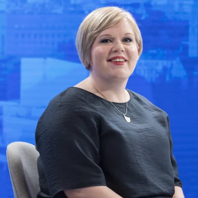 Annika Saarikko sitter på en stol vid ett bord. Framför henne finns ett glas vatten och en mikrofon. Bakgrunden består av en elektronisk duk som är blå.