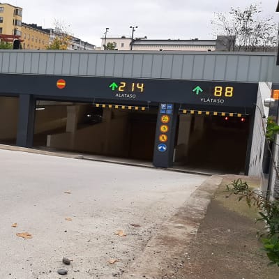 Valkoinen henkilöauto ajaa pysäköintihallin luiskaa pitkin kohti sisäänkäyntejä. Sisäänkäyntien yllä on valokirjaimin ilmoitettu, kuinka monta paikkaa on vapaana.