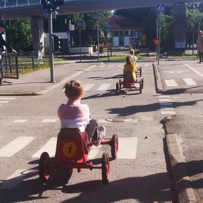Barn på röd trampbil i övningslandskap för trafik, med övergångsställen, korsningar och trafikljus.