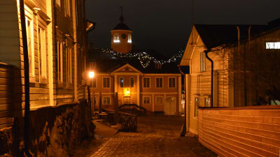 Vy mot rådhuset i Borgå. Gamla trähus kantar en kullerstensgata som leder ut på ett kullerstensbelagt torg. På andra sidan torget står ett stort hus med ett upplyst klocktorn. Det är mörkt men vyn lyses upp av lampor och julljus.