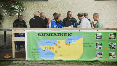 Grupp med män dricker kaffe och pratar på en terrass. Terassräcket pryds av stor karta över byn Sumiais.