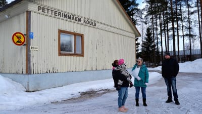 Tre personer står intill ett hus och pratar. Det är vinter. På husväggen står det Petterinmäen koulu.