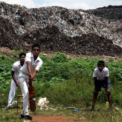 Kolonnawan jätevuori romahti myös vuonna 2012, jolloin yli 50 taloa jäi jätemassan alle rankkasateiden seurauksena. Kuvassa etualalla valkoisiin pukeutuneet nuoret pojat pelaavat krikettiä. Taustalla näkyy karmaiseva, haiseva jätevuori. 