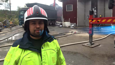 Brandman Terence Sam står framför en industrihall där eftersläckningsarbete pågår.