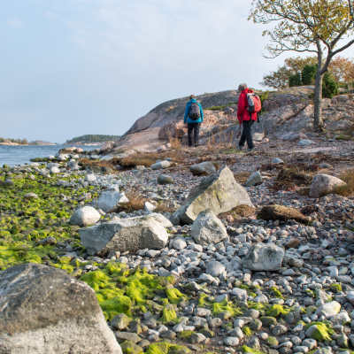 Två personer med jackor och ryggsäckar vandrar längs en stenig strand på en ö i Skärgårdshavets nationalpark.