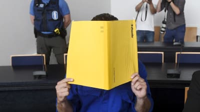 En person sitter och håller en gul mapp framför ansiktet i en rättegångssal. En polis och två fotografer står i bakgrunden av bilden.