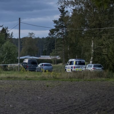 Ett trähus och parkerade bildar syns från en åker. Bland bilarna finns också en polisbil.