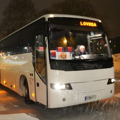 En vit buss står på en parkeringsplats. Inne i den annars tomma bussen sitter en äldre man som är chaufför. Uppe på bussen står Loviisa.