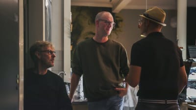 Man med glasögon står i mörklagt kafé och pratar med en man med hatt.