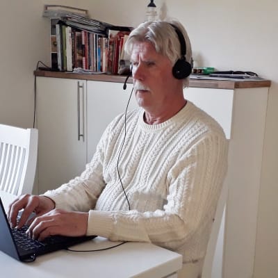 En man i övre medelåldern har headset på huvudet och sitter vid sitt köksbord framför sin laptop och skriver.
