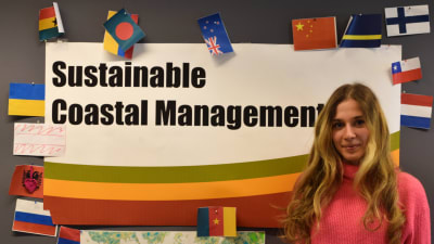 En ung kvinna framför en poster där det står Sustainable Coastal Management. Runt postern finns det flaggor från flera olika länder.