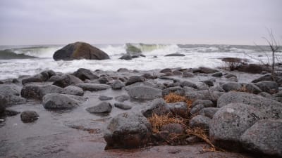 En strand med stora släta stenar. Stora vågor slår mot stenarna och himlen är grå.