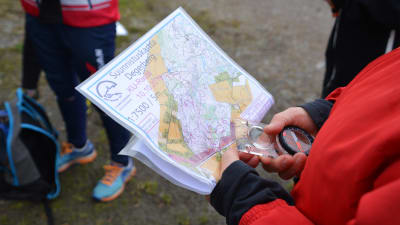 Närbild på en orienteringskarta och en kompass som någon i röd jacka håller i.