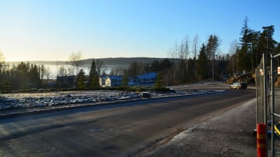 En asfalterad väg leder ner mot några trähus. Nära fotografen, i högra hörnet av bilden, finns ett stängsel runt en byggplats. I fjärran, bakom trähusen, syns Borgå å.