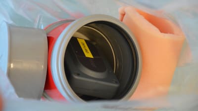 I mitten av bilden synns en fyrkantig svart dosa som det står önyland på. Dosan ligger inuti en grå plastburk med röd tejp på sidan. Till vänster om plastburken ligger ett grått lock och till höger en skumgummibit.