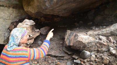 En medelålders man i randig tröja och duk om huvudet står framför en låg grottöppning. Han pekar inåt grottan.