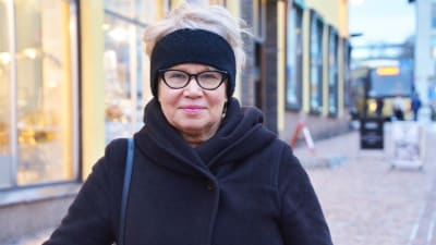 En medelålders kvinna i svart kappa och svart pannband står utanför ett gult hus i Gamla stan i Borgå. Hon tittar in i kameran och ler lite.
