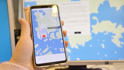 En hand håller i en smarttelefon med en karta på skärmen. Bakom handen och telefonen syns en dator med en karta på skärmen.