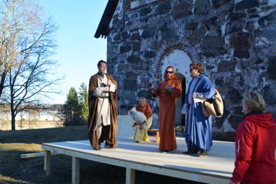 Fyra personer klädda i långa kåpor och kvinnorna i huvuddukar står på en scen uppbyggd framför Sibbo gamla kyrka.