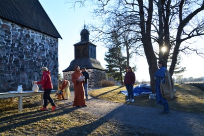 Sex personer står utanför Sibbo gamla kyrka. Vissa av dem är klädda i gammaldags kläder, långa kåpor och huvuddukar.