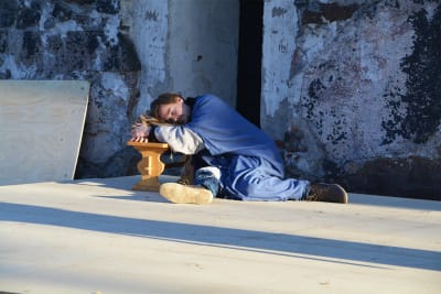 En man iklädd lång kåpa och blå mantel halvligger på en scen. Han har huvudet på en säck lutad mot en bänk och ser ut att sova.