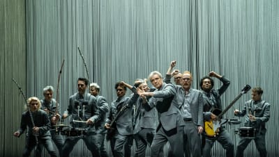 David Byrne ja orkesteri lavalla, harmaissa puvuissa, kuva konserttielokuvasta David Byrne's American Utopia.