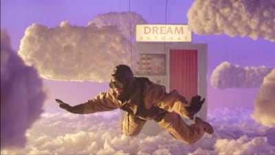 Vanhan ajan lentäjän asuun pukeutunut hahmo leijuu pilvien päällä studiolavasteissa, taustalla näkyy Dream Automat -kuvauskoppi.