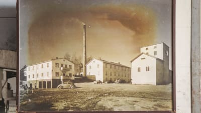 Svartvitt foto på gamla fabriksbyggnader, en fabrikspipa och 1950-talsbilar.