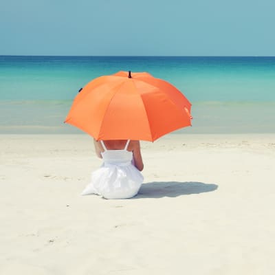 Kvinna sitter under ett parasoll på en sandstrand. 