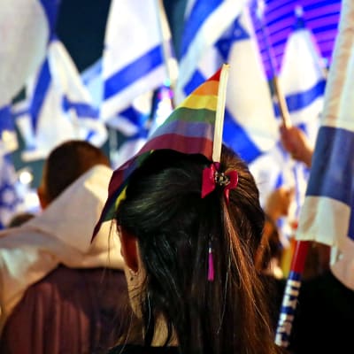 En kvinna med regnbågsflagga i håret fotograferad bakifrån. Flera israeliska flaggor runtomkring.
