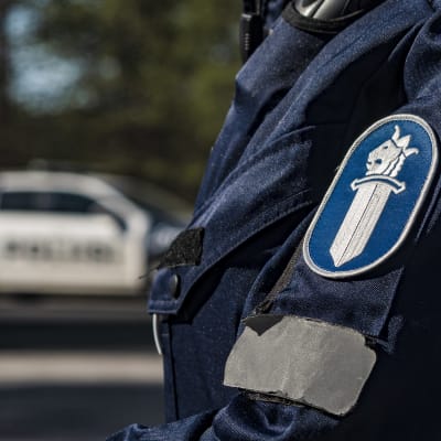 Närbild av polisens logo på en polisuniform. I bakgrunden syns en polisbil.