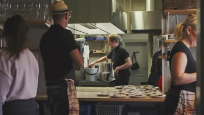 Restaurangkök, kock rör om i gryta, i förgrunden färdiga portioner på bord.