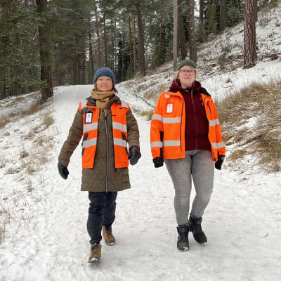 Oranssiseen huomioliiviin pukeutunut Kaarina Aulin ja huomiotakkiin pukeutunut Leena Väisänen kävelevät lumisen metsäpolun mäkeä alas kohti kameraa.