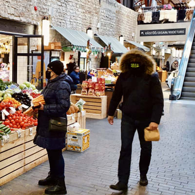 Grönsaksförsäljning på inomhusmarknaden Balti Jaama Turg i Tallinn. De fyra personerna på bilden använder munskydd på olika sätt - från inget till munskydd och jackans huva tätt dragna över ansiktet.