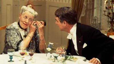 Frakkipukuinen mies puhuttelee ruokapöydän ääressä vanhaa rouvaa, joka käyttää kuulotorvea.
