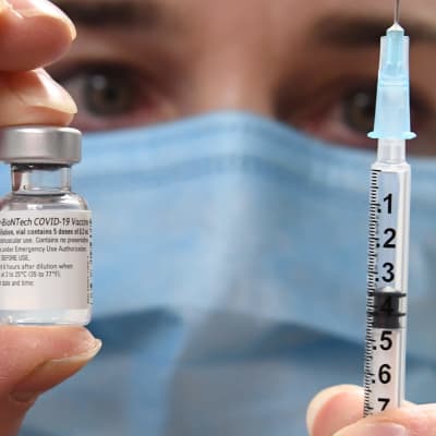 I bakgrunden syns ett ansikte med munskydd. Kameran fokuserar på en liten glasflaska med coronavaccin och en spruta personen håller i.