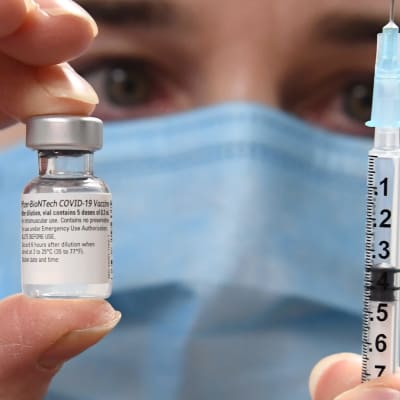 I bakgrunden syns ett ansikte med munskydd. Kameran fokuserar på en liten glasflaska med coronavaccin och en spruta personen håller i.