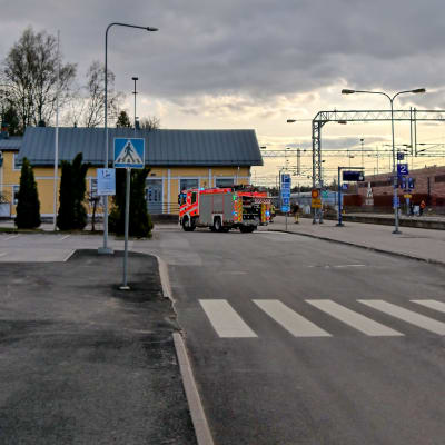 En brandbil står parkerad utanför en gul träbyggnad som är en järnvägsstation. Till höger löper järnvägsspåren.