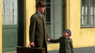 hattupäinen mies laukku kädessään pitää lakkipäistä pikkupoikaa kädestä keltaisen talon ulko-oven luona. Kuva sarjasta Matador.