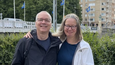 En man med glasögon, ljust kort hår och blå jacka, och en kvinna med glasögon, axellångt ljust hår och en vit jacka håller om varandra på en strandgata, med en bro i bakgrunden där Åbo stads blå stadsvapen flaggar.