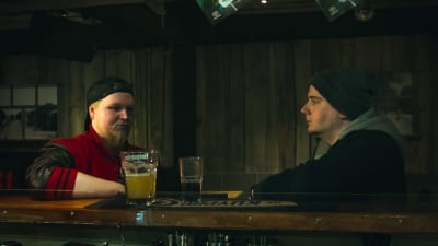 Kaksi nuortamiestä istuvat totisina hämärässä pubissa tuopit edessään pöydällä. Vasemmanpuoleisella on päässään lippalakki, oikeanpuoleisella pipo.