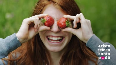 Hymyilevä tyttö pitää sormillaan kahta mansikkaa silmiensä edessä.