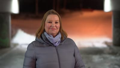 En kvinna i vinterrock ser mot kameran och småler. Det är kväll.