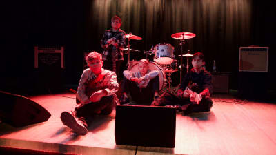 Fyra bandmedlemmar sitter på golvet med sina instrument och tittar in i kameran.