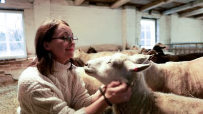 Mörkhårig kvinna med glasögon ler och smeker ett blundande får. 