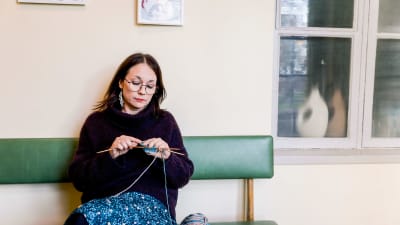 Mörkhårig kvinna i ylletröja och blommig klänning sitter på en bänk i ett väntrum och stickar.