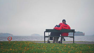 Iäkäs pariskunta selin kuvaan istumassa puiston penkillä järven rannassa