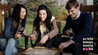 Kolme aikuista pelaa yhdessä Scrabblea terassilla.